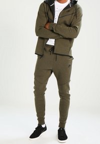 Olivegrön Nike hoodie och joggerset med svarta detaljer, med dragkedja fram, ribbade ärmslut och slim-fit design, tillsammans med svarta sneakers.