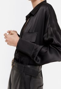 Blusa nera in satin con apertura a bottoni, taschino e maniche ampie. Abbinata a pantaloni in pelle nera a vita alta e una cintura.
