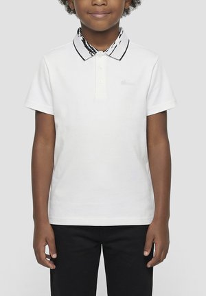 Niño con una camiseta tipo polo blanca de manga corta con un cuello estampado en blanco y negro y pantalones negros sobre un fondo liso.