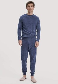 Dunkelblaues Loungewear-Set mit langärmligem Oberteil und schmal zulaufenden Hosen, beide aus weichem strukturiertem Stoff. Modell trägt helle Hausschuhe.