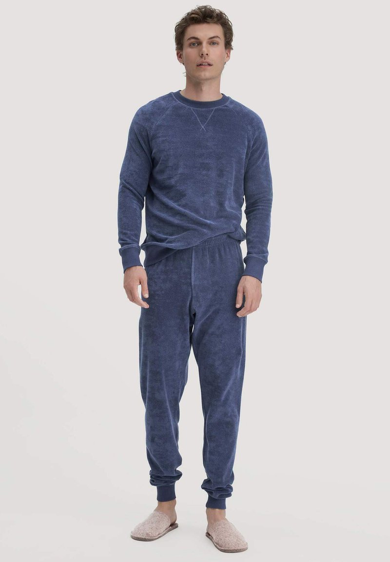 Dunkelblaues Loungewear-Set mit langärmligem Oberteil und schmal zulaufenden Hosen, beide aus weichem strukturiertem Stoff. Modell trägt helle Hausschuhe.