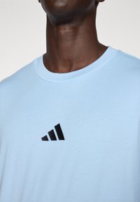 T-shirt de algodão azul claro com gola redonda e logótipo bordado em preto com três riscas no peito. Textura suave, design casual.