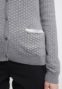 Grauer Strickcardigan mit strukturiertem Punktmuster, Knopfverschluss und einer weißen aufgesetzten Tasche auf der linken Seite.