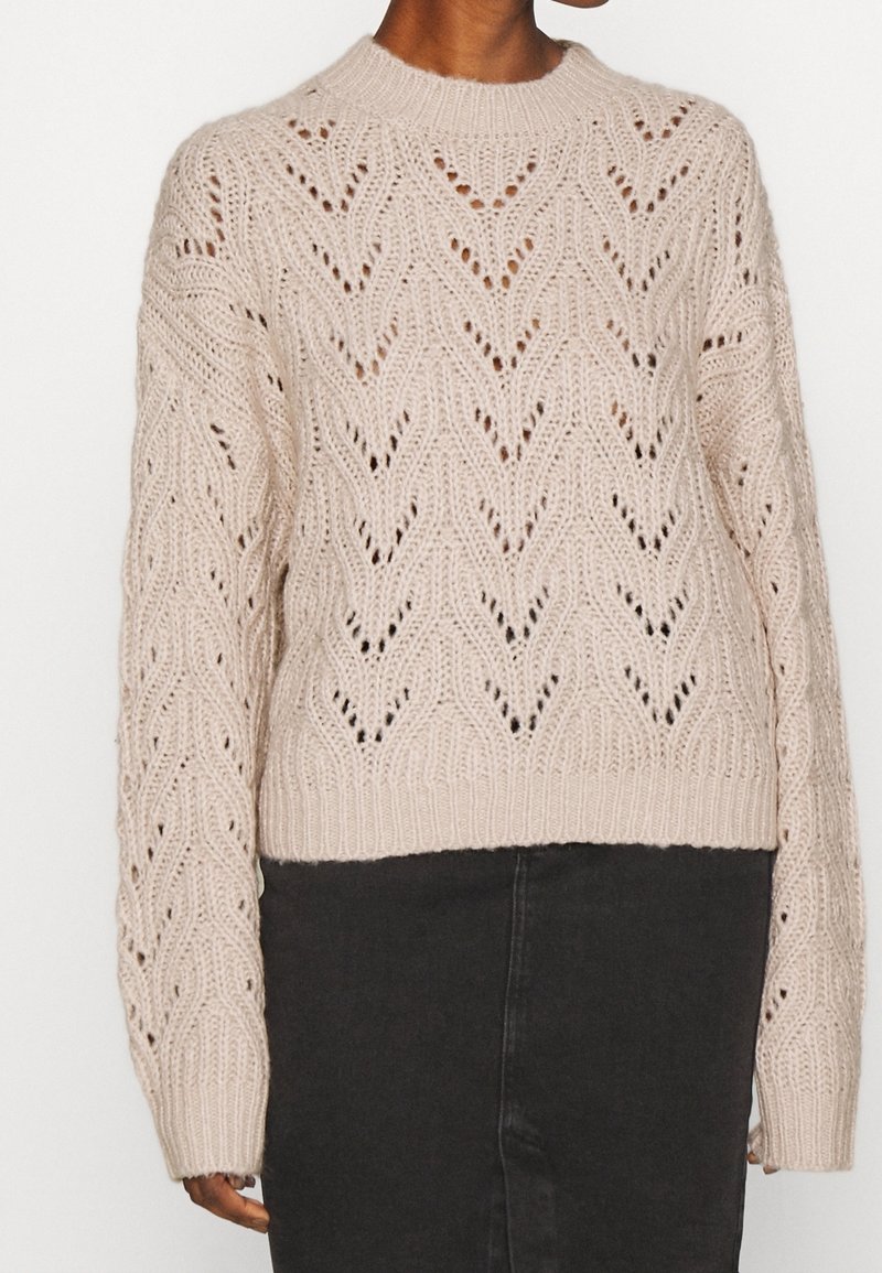 Personne portant un pull en maille texturée, beige clair, à manches longues avec un motif chevron, assorti à une jupe noire taille haute.