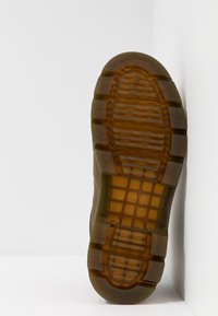 Suela de goma translúcida marrón con patrón de textura, mostrada en posición vertical contra un fondo de pared y suelo blancos.