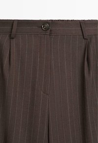 Gros plan d'un pantalon marron à fines rayures avec passants de ceinture, plis, fermeture à boutons et fines rayures blanches verticales.