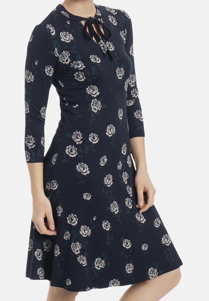 Femme portant une robe bleu marine avec des motifs de roses blanches et des manches trois-quarts, avec un col en forme de trou de serrure noué.