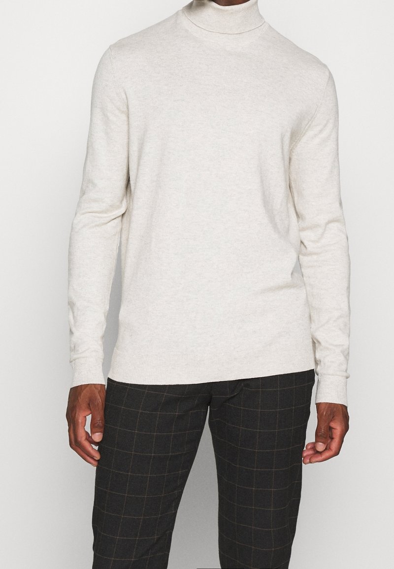 Hellbeige Rollkragensweater aus weichem Strickmaterial mit langen Ärmeln und gerippten Bündchen, kombiniert mit dunklen Karohosen.