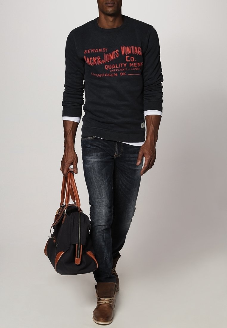 Felpa grigia scura con testo stampato rosso, jeans in denim blu, stivali marroni e una borsa nera con accenti marroni. Abbigliamento casual con texture stratificate.