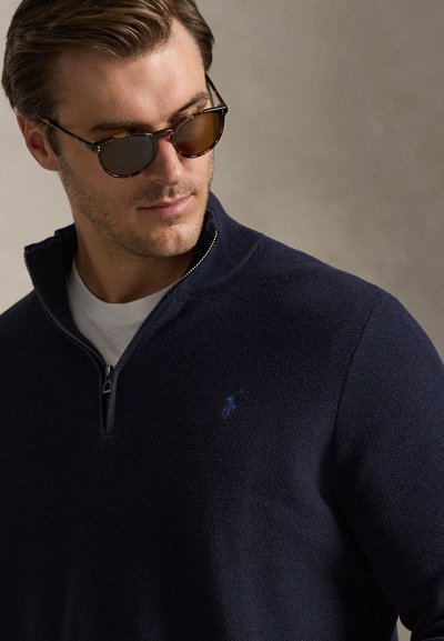 Homme portant des lunettes de soleil écaille de tortue et un pull bleu marine à fermeture éclair sur un quart avec un petit logo brodé, regardant vers le bas et légèrement sur le côté.