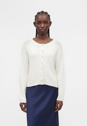 Γυναίκα με πλεξούδες στα μαλλιά, φορώντας λευκή πλεκτή ζακέτα με κουμπιά και μια σκούρα μπλε φούστα, στέκεται μπροστά σε ένα απλό λευκό φόντο.