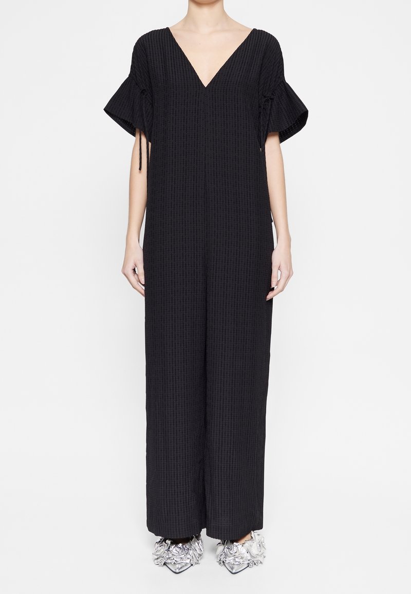 Femme portant une combinaison ample noire texturée avec des manches courtes à volants et des chaussures argentées pointues ornées de motifs floraux.