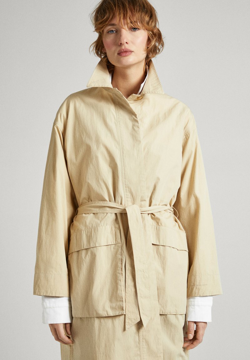 Veste beige à ceinture avec col montant, deux grandes poches avant, coupe loose et manches longues sur une chemise blanche. Texture lisse.
