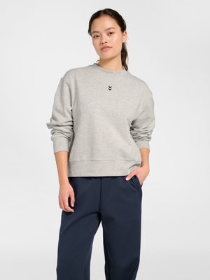 Ung kvinde med langt mørkt hår i hestehale iført lys grå sweatshirt og mørkeblå bukser, stående med den ene hånd i lommen.