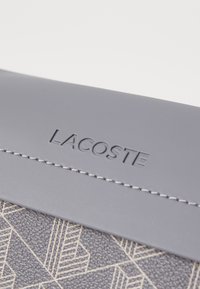 Harmaa keinonahkainen pussi, jossa on geometrinen kuvio, kohokuvioitu "LACOSTE"-logo ja kontrastivärinen ompelu yläreunassa.