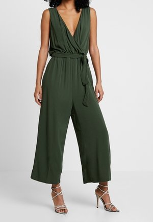 Femme portant une combinaison sans manches vert olive à décolleté en V profond, avec une taille nouée et un pantalon large raccourci, associée à des sandales à talons hauts à lanières.