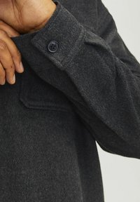 Dark grey wool shirt with a buttoned cuff and a textured finish. Features a visible pocket detail on the sleeve.