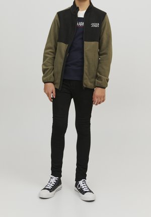 Osoba ubrana w oliwkowo-czarną polarową kurtkę zapinaną na zamek marki Jack & Jones, granatowy t-shirt z grafiką, czarne obcisłe dżinsy oraz czarne wysokie trampki z białymi sznurowadłami.