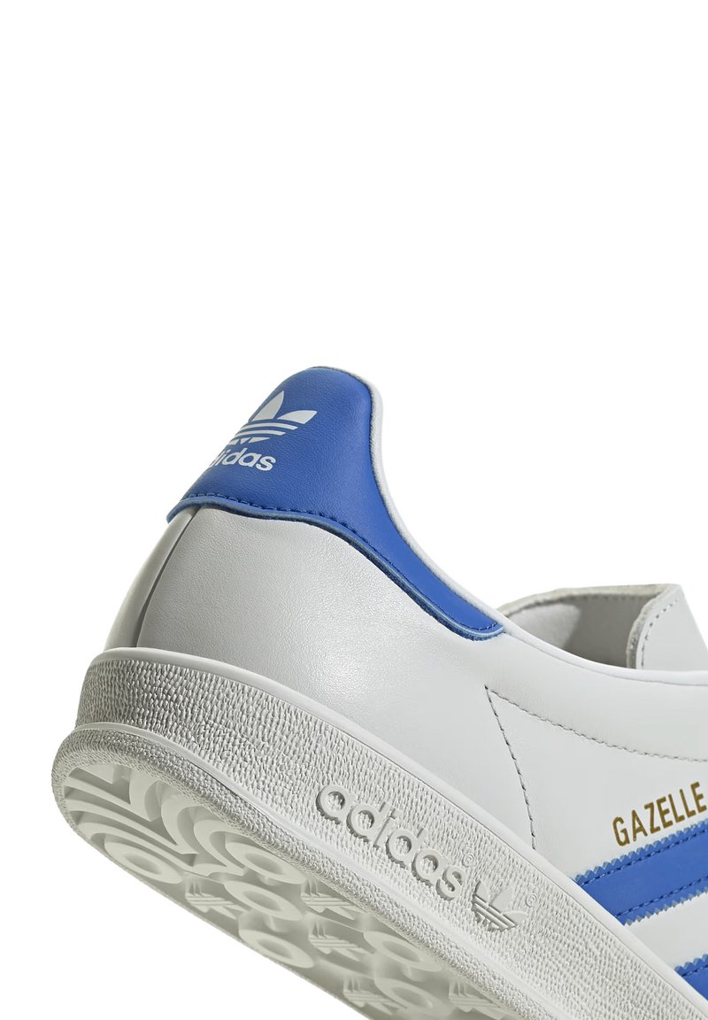 Baskets blanches avec des accents bleus et l'inscription dorée "Gazelle", dotées d'une semelle texturée et du logo Adidas sur le talon.