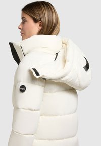 Chaqueta acolchada blanca con un alto cuello y capucha desmontable. Presenta paneles cosidos, un logo en el lateral y acentos en negro. Textura suave.