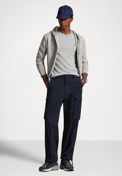 Ανοιχτό γκρι φούτερ με φερμουάρ πάνω από ανοιχτό γκρι μπλουζάκι, σκούρο μπλε παντελόνι cargo και σκούρα αθλητικά παπούτσια. Ναυτικό καπέλο με κόκκινες λεπτομέρειες λογότυπου.