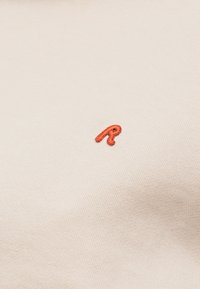 Μικρό κόκκινο κεντητό γράμμα "R" σε ανοιχτό μπεζ ύφασμα με υφή από λεπτές διαγώνιες γραμμές.