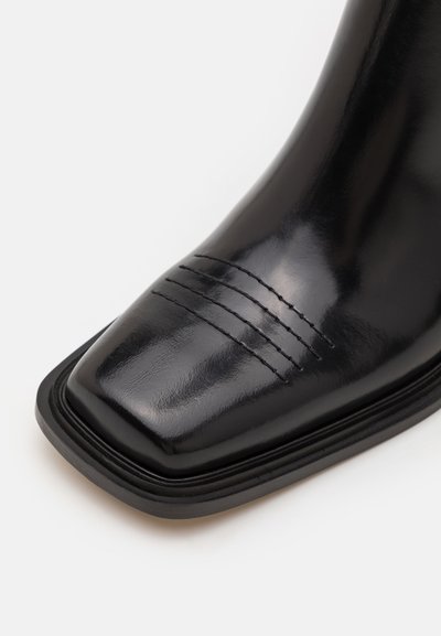 Black leather ankle boot with a square toe, featuring three horizontal stitching lines on the toe cap and a raised black rubber sole.