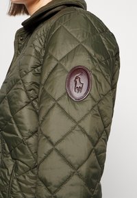 Olivgrön vadderad jacka med en strukturerad yta, med ett brunt läderemblem på vänster axel och en avtagbar huva.