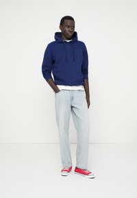 Haut à capuche bleu marine avec broderie de logo, associé à un jean droit bleu clair et des baskets basses rouges. T-shirt blanc uni en dessous.