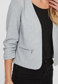 Šedý strukturovaný blazer s kratším střihem, dvěmi předními kapsami a zipovým zapínáním. Nosený přes černý top s vyhrnutými rukávy.