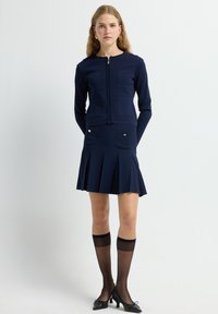 Chaqueta de punto texturizado en azul marino con cierre de cremallera, combinada con una falda plisada a juego que cuenta con bolsillos frontales y detalles en tono dorado.