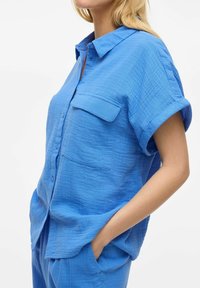 Camicia a maniche corte con bottoni in un tessuto blu testurizzato, dotata di taschino sul petto e polsini arrotolati.