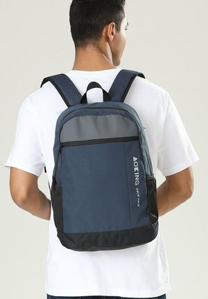 Mann trägt weißes T-Shirt und einen marineblauen und grauen Rucksack mit schwarzen Gurten, steht mit dem Rücken zur Kamera vor einem schlichten, hellen Hintergrund.
