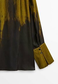 Chemise à manches longues noir et vert foncé en tissu texturé, avec des poignets vert clair contrastants et des détails de boutons noirs.