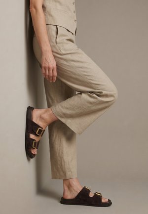 Personne portant un pantalon et un gilet en lin beige, appuyée contre un mur, avec une jambe pliée et des sandales marron foncé à boucle aux pieds.