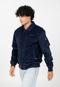 Marineblauwe corduroy bomberjack met een ritssluiting aan de voorkant, kraag en een gestructureerde afwerking. Voorzien van zijzakken en een patch op de linkerborst.