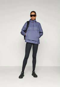 Chaqueta impermeable ligera de color púrpura con media cremallera, capucha y bolsillo en el pecho; combinada con leggings negros y zapatillas deportivas. Lleva puestas gafas de sol oscuras.