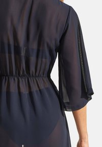 Couverture transparente marine avec taille élastique, manches trois-quarts et tissu doux et fluide. Présente un éclat subtil et une texture légère.