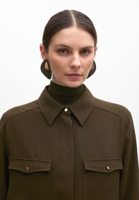Blusa marrone con colletto e due tasche sul petto, dotata di hardware dorato. Indossata con un dolcevita a coste verde scuro e grandi orecchini neri.