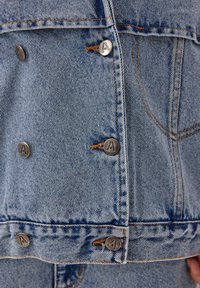 Zbliżenie na jasnoniebieską kurtkę dżinsową z przodu, z metalowymi guzikami grawerowanymi w trójkątny logo i pomarańczowymi detalami stębnowania.