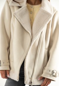 Donna che indossa una giacca in shearling beige con fibbie argentate e zip sopra un maglione giallo abbottonato e pantaloni scuri.