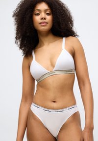 Baltas medvilninių liemenėlė su reguliuojamomis petnešėlėmis, su smėlio spalvos Calvin Klein juosta. Atitinkamos baltos thong kelnaitės su juodos spalvos logotipu.