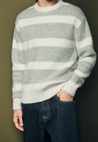 Gestreifter Pullover in Hellgrün und Weiß, gestrickte Textur, Rundhalsausschnitt, gerippte Bündchen und Saum, getragen mit dunklen Jeans.