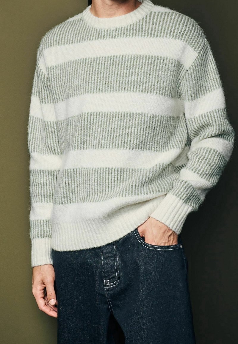 Gestreifter Pullover in Hellgrün und Weiß, gestrickte Textur, Rundhalsausschnitt, gerippte Bündchen und Saum, getragen mit dunklen Jeans.