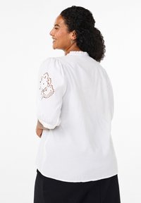 Vit blus med puffärmar som har dekorativ spetsbroderi på ärmarna och en volangkrage, tillverkad av lättviktsmaterial.