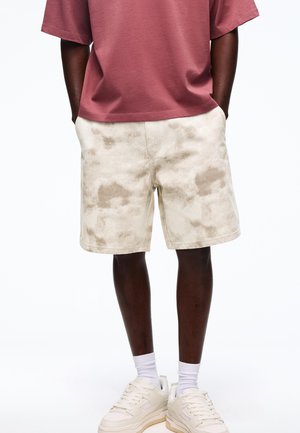Mand iført beige tie-dye shorts, hvide sokker, hvide sneakers og en løstsiddende t-shirt i rustfarve med hænderne i lommerne.