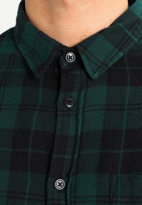 Zeleno-černý kostkovaný košil s knoflíkovým límcem, s výraznou texturou látky a černými knoflíky. Viditelné švy a detail kapsy.