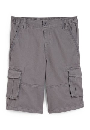 Barn iført en sort PlayStation-t-shirt, grå cargobukser, hvide sokker med sorte striber og sorte og hvide sneakers, stående mod en hvid væg.