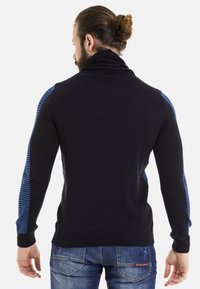 Dunkelblauer, gestrickter Pullover mit blauen gestreiften Seiteneinsätzen, hohem Kragen, gerippten Bündchen und Saum. Getragen mit blauen Denim-Jeans, von hinten gezeigt.