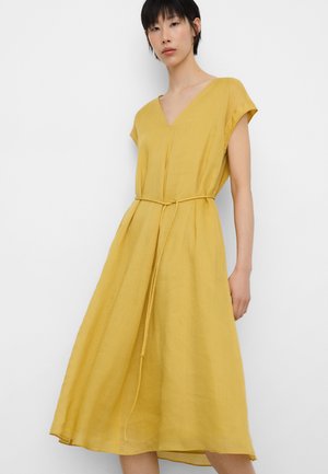 Kobieta w luźnej, bezrękawnikowej sukience w kolorze musztardowej żółci z dekoltem w kształcie litery V i cienkim paskiem przewiązanym w talii, stojąca na tle białego tła.